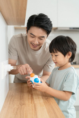 Adult male supervising a young boy playing with a colorful electronic robot toy at a wooden table in a kitchen, illustrating parental supervision with electrical toys.